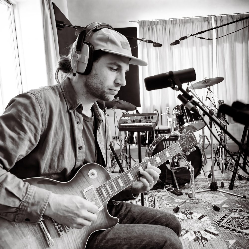 Black and white photo of a musician playing electric guitar with a drum kit behind